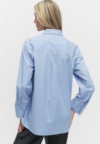 Kobieta z blond włosami, nosząca jasno-niebieską, paskowaną koszulę z długim rękawem, ukazana z tyłu, stojąca na tle gładkiej, białej ściany.