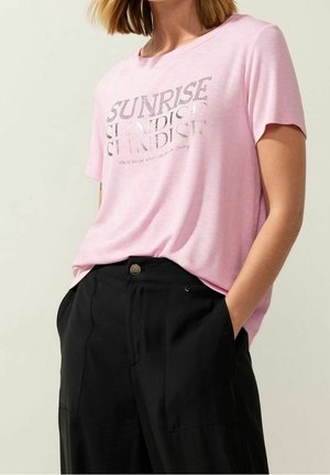 Kvinde iført en lyserød T-shirt med teksten "SUNRISE SUNSET" og sorte bukser med hænderne i lommerne, står mod en lys baggrund.