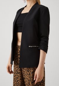Černé sako s detailem předního zipu, dlouhými rukávy a hladkou strukturou, spojené s kalhotami se zvířecím leopardím vzorem.