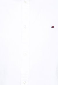 Λευκό πουκάμισο με κουμπιά και υφή από κλωστοϋφαντουργικό υλικό, που διαθέτει ένα μικρό κεντημένο λογότυπο σε κόκκινο, λευκό και μπλε χρώμα στο αριστερό μέρος του στήθους.