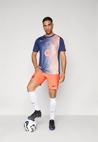 Mees, kes kannab tumesinist ja oranži FC Barcelona jalpallivormi, seisab üks jalg jalpallil tasase tausta ees.