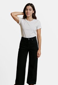 Witte katoenen t-shirt gecombineerd met hoge zwarte wijde jeans. De jeans hebben een knoopsluiting aan de voorkant en zijzakken. Effen achtergrond.