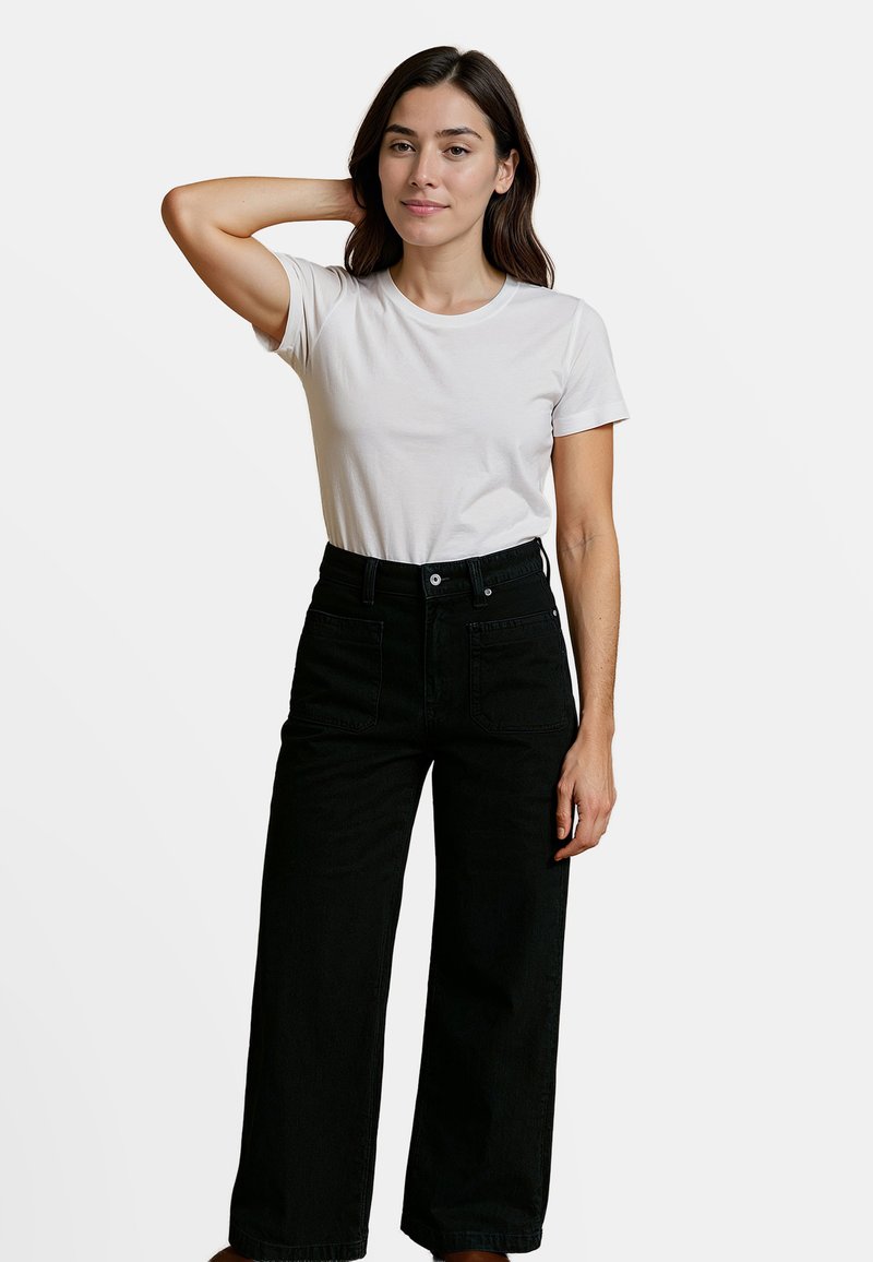 Witte katoenen t-shirt gecombineerd met hoge zwarte wijde jeans. De jeans hebben een knoopsluiting aan de voorkant en zijzakken. Effen achtergrond.