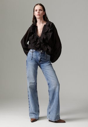 Une femme porte un chemisier noir à volants, un jean bleu à jambes larges et des chaussures marron devant un fond de studio gris uni.