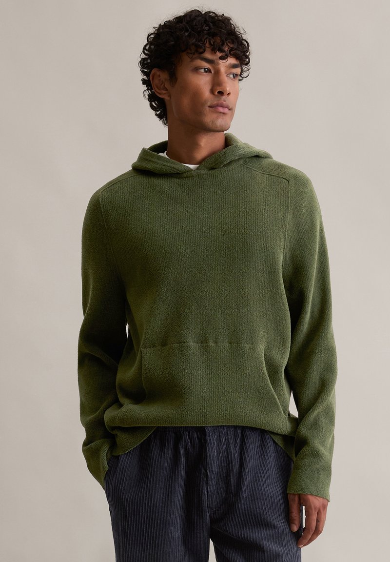 Πράσινη υφασμάτινη κουκούλα με μπροστινή τσέπη και κουκούλα, σε συνδυασμό με σκούρους ριγέ παντελόνες. Το ύφασμα φαίνεται απαλό και έχει χαλαρή εφαρμογή.