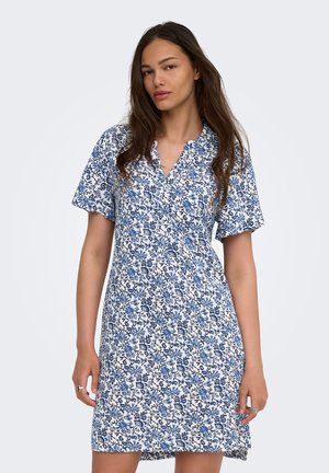 Kvinde med langt brunt hår iført en hvid knælængde kjole med blå blomsterprint og korte ærmer mod en ensfarvet baggrund.