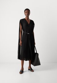 Black pleated dress with a V-neck, cape sleeves, and waist belt. Model holds a black tote bag, paired with black open-toe mules.