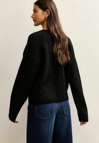 Jeune femme aux longs cheveux bruns, portant un pull noir et un jean bleu, debout de profil contre un mur beige uni.
