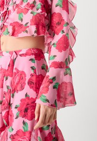 Completo due pezzi floreale rosa realizzato in tessuto trasparente, caratterizzato da audaci rose rosse e foglie verdi, con dettagli arricciati sulle maniche e sull'orlo.