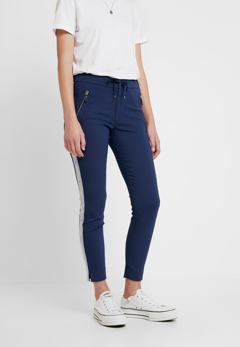 Marina blå joggerbyxor med dragkedjefickor, dragsko i midjan och kontrasterande silverfärgade sidoströmmar; tillverkade av elastiskt material med slim fit.