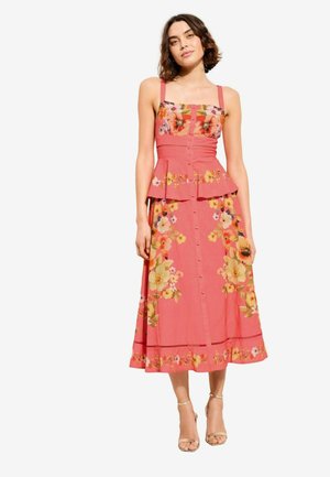 Žena v koralových midi šatách bez rukávov s kvetinovými potlačami, s gombíkmi, so zavinovacím pásom v páse a béžovými členkovými opätkami stojaca na bielom pozadí.
