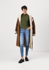 Άνθρωπος που φοράει ένα μακρύ καφέ παλτό από γούνινη επένδυση πάνω από ένα ελαιόλαδο ριγέ μπλουζάκι, ανοιχτό μπλε στενά τζιν και μαύρα παπούτσια με φούντες, στέκεται με το ένα χέρι στην τσέπη.