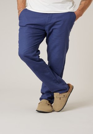 Homme portant un pantalon bleu et des chaussures beige à enfiler, debout avec les jambes croisées et les mains dans les poches, devant un fond uni.