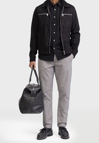 Schwarze Wildlederjacke mit silbernen Reißverschlüssen, schwarzes Hemd mit Knöpfen, graue karierte Hose und eine schwarze Lederreisetasche. Klassische Schnürschuhe.