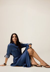 Donna con lunghi capelli scuri seduta a terra, che indossa un vestito blu con cintura e maniche arrotolate, e sandali neri con tacco basso, su uno sfondo neutro.