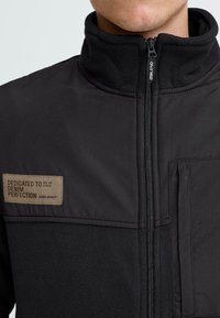 Crni zip-up jakna s flis ovratom, džepom na prsima i tkanim pecatom s natpisom "POSVEĆENO SAVRŠENSTVU DENIMA."