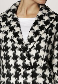 Schwarz-weiß karierter Mantel im Hahnentrittmuster aus weichem Material, mit einem Schlitzkragen und einer doppelt geknöpften Vorderseite.