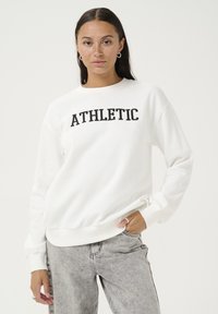 White fleece sweatshirt with "ATHLETIC" printed in bold black letters; features crew neck and ribbed cuffs, paired with gray jeans.