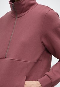 Gros plan sur une personne portant un pull à fermeture éclair quart de zip rose poussiére avec des poignets côtelés et une poche kangourou, la main reposant à l'intérieur de la poche.