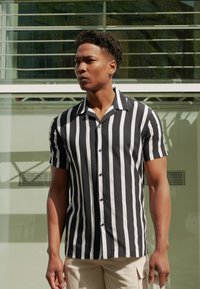 Chemise à manches courtes avec des rayures verticales noires et blanches, fabriquée en tissu léger, dotée d'une fermeture à boutons sur le devant et d'un col pointu.