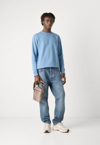 Sudadera azul clara con puños acanalados y cuello redondo, combinada con jeans azules holgados. Lleva un pequeño bolso de hombro beige con logotipo.