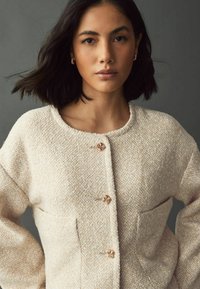 Beige textured cardigan with a round neckline, two front pockets, and decorative knot-style buttons. Soft fabric with a subtle pattern.