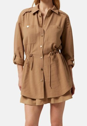 Femme portant une chemise à boutons brun clair avec taille à cordon de serrage, manches retroussées et poches poitrine à l'avant avec boutons en métal.