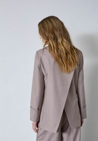 Svetlohnedé sako na mieru s unikátnym otvoreným zadným dizajnom. Má dlhé rukávy a čisté línie, vyrobené z hladkej, štruktúrovanej látky.