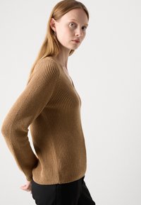 Pull à col en V en texture côtelée, fabriqué à partir d'un matériau marron clair. Il présente des manches longues et une silhouette ajustée.