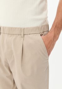 Beige, lahki hlače z elastičnim pasom, stranskimi žepi in sprednjimi gubami; tkanina se zdi gladka z rahlim sijajem.