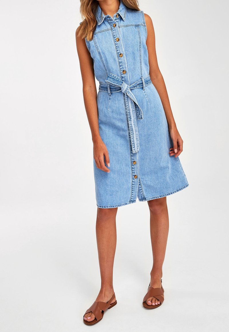 Světle modré džínové šaty košilového střihu bez rukávů, s knoflíkovým zapínáním a látkovým páskem. Mají délku ke kolenům a volný střih.
