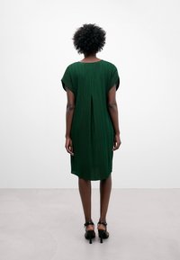 Kvinna med kort, lockigt hår, klädd i en knälång, mörkgrön, veckad klänning och svarta högklackade sandaler, stående mot en enfärgad vit bakgrund.