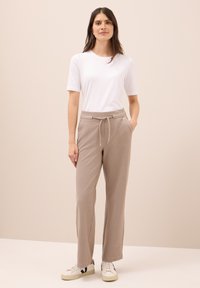 Een vrouw staat tegen een beige achtergrond en draagt een wit T-shirt met korte mouwen, beige broek met trekkoord en witte sneakers met zwarte accenten.