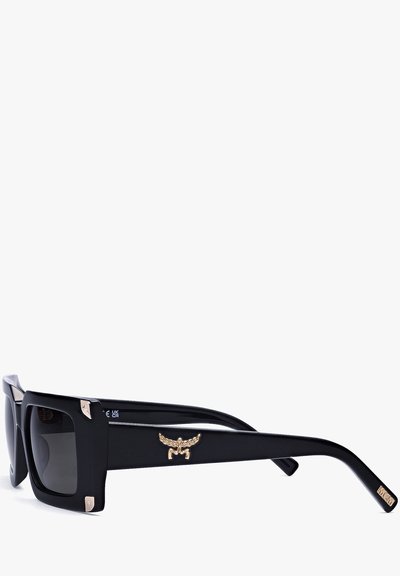 Zwarte oversized zonnebril met vierkante glazen, voorzien van gouden metalen accenten en een gevleugeld embleem op de brilpoot. Gladde plastic montuur.