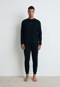 Cămașă neagră cu mâneci lungi și pantaloni jogger asortați, confecționați dintr-un material moale, având un mic logo roșu pe pieptul cămășii.