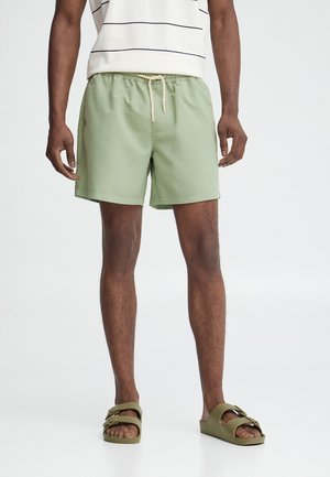 Hombre que lleva pantalones cortos verde claro con cordón, camiseta sin mangas de rayas blancas y negras, y sandalias verdes oliva sin pulsera.