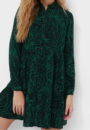 Vrouw draagt een groene en zwarte abstracte zebra gestreepte jurk met lange mouwen, knopen aan de voorkant en een gelaagde rok.