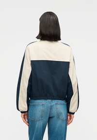 Μπουφάν αμάνικο σε μπλε ναυτικό και εκρού με ελαστικές μανσέτες και τελείωμα, με σχέδιο σε χρωματιστά μπλοκ. Το υλικό έχει ομαλή υφή.