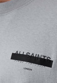 Camiseta de algodón gris con una raya horizontal negra que presenta el texto en relieve "ALLSAINTS" y caracteres japoneses, junto con "LONDRES" impreso abajo.