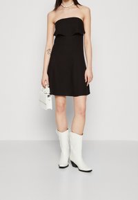 Robe noire sans bretelles avec un haut à volants, associée à des bottes de cowboy blanches ornées et à un petit sac à main blanc. Texture de tissu lisse.