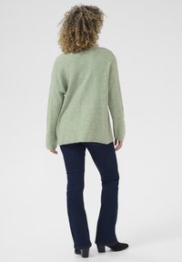 Pull en maille vert clair avec une coupe ample, des manches longues et un ourlet asymétrique, associé à un jean évasé bleu foncé et des talons noirs.