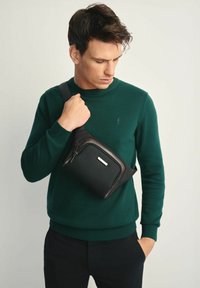 Czarna torba na ramię wykonana z teksturowanego materiału, z przednią kieszenią na zamek i gładkim brązowym akcentem. Regulowany pasek dla wygody.