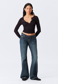 Zwarte langärmelige top met een drukknopdetail, gecombineerd met donkerblauwe flared jeans. De outfit heeft een casual pasvorm en een soepele textuur.