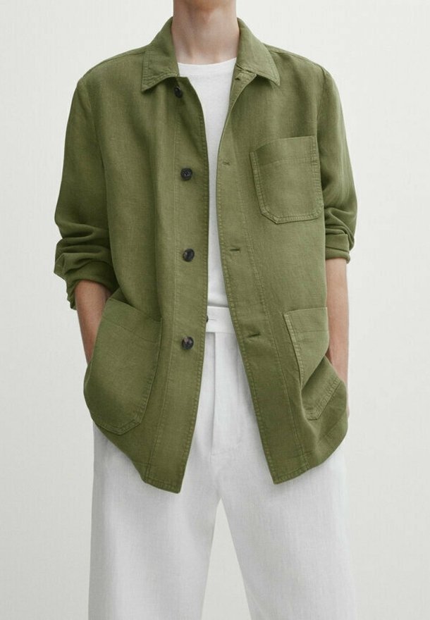 Personne portant une veste boutonnée vert olive avec trois poches avant et des manches retroussées par-dessus une chemise blanche et un pantalon blanc.