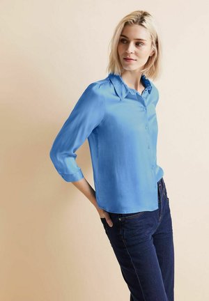 Camisa de botones azul clara con cuello y mangas tres cuartos, hecha de una tela suave, combinada con unos vaqueros de denim oscuro.