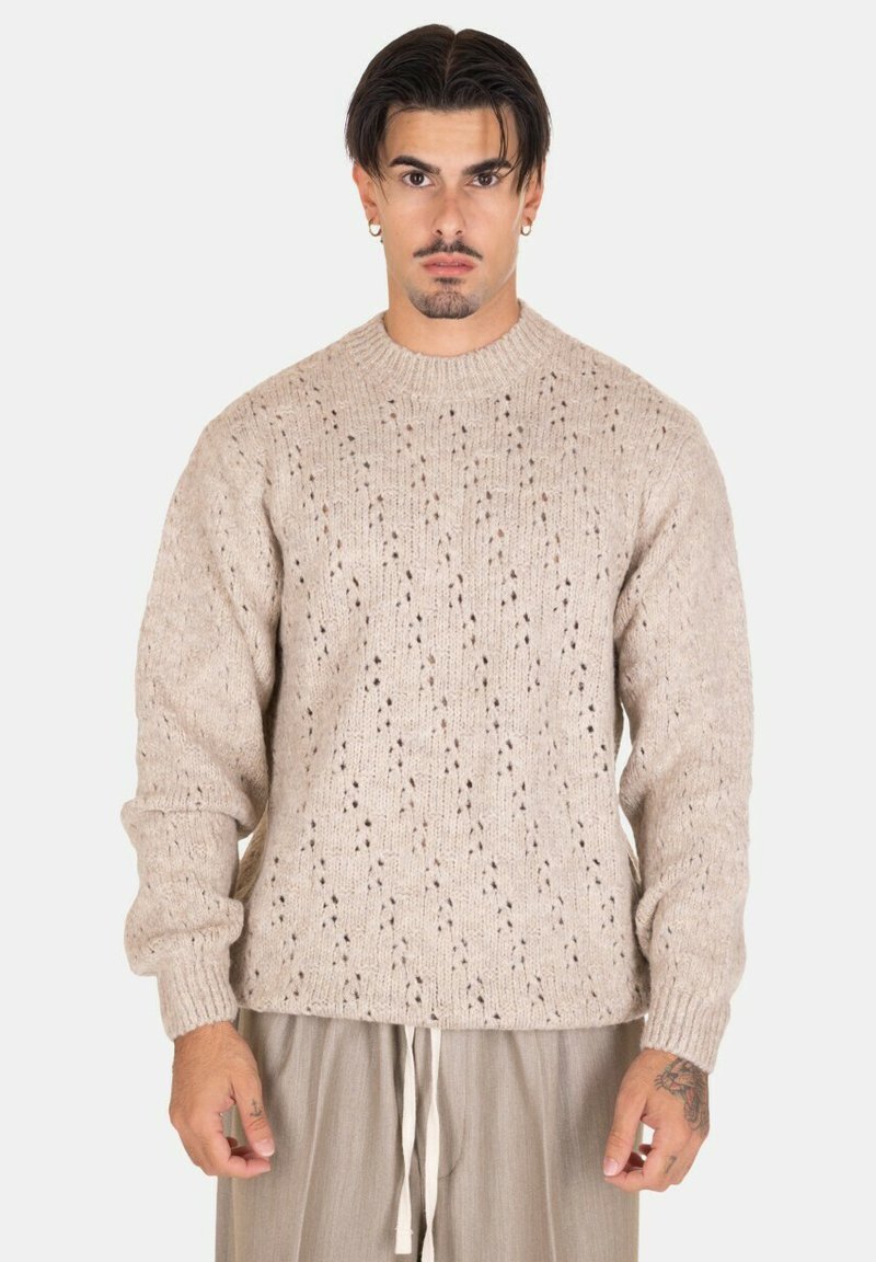 Maglione lavorato a maglia beige con un motivo strutturato, collo rotondo e polsini a costine. Indossato sopra pantaloni chiari e larghi.