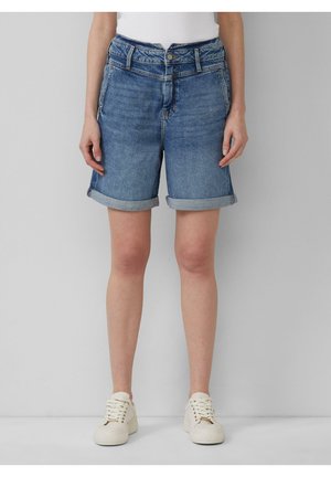 Denim shorts met opgerolde boorden gedragen door een staand persoon, gecombineerd met witte sneakers en een ingestopt wit topje.