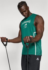 Zaļa bezpiedurkņu Adidas treniņu kreklā ar baltu logo, kombinēta ar melniem šortiem. Modelis tur vingrošanas lentas ar strukturētām rokturiem.