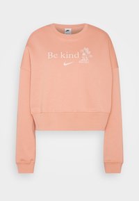 Ljust persikofärgad kort sweatshirt med långa ärmar, vit text "Var snäll", Nike-logga och linjeteckning av en sko med blommor som växer ur den.