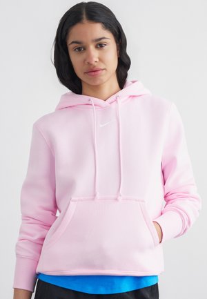 Jeune femme aux cheveux noirs mi-longs portant un sweat à capuche Nike rose clair par-dessus une chemise bleue, les mains dans les poches, face à l'avant.